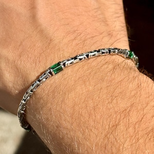 Könnte beinhalten: Ein silbernes Armband mit Kettenmuster, verziert mit rechteckigen smaragdgrünen Steinen. Das Armband wird am Handgelenk getragen und zeigt das detaillierte Muster und den Kontrast zwischen Silber und grünen Edelsteinen.