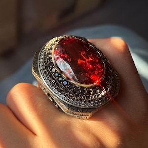 May include: A detailed silver ring with a large, oval-shaped, deep red gemstone. The ring is embellished with smaller, clear and dark gemstones around the central stone, set within an intricate metal design.