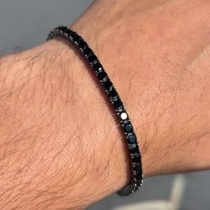May include: A black gemstone bracelet. The bracelet is made of small, round, black gemstones set closely together. The bracelet is worn on a wrist, and the background is blurred.