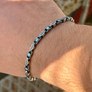 May include: A silver bracelet with a chain-link design, featuring small, round, light blue stones. The bracelet is worn on a wrist, with the skin tone visible in the background. The bracelet's links have an ornate pattern.