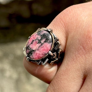 May include: A silver ring with a pink and black oval gemstone set in a detailed silver band.