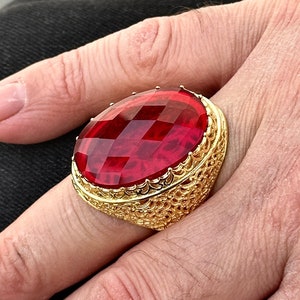 May include: A gold ring with a large, red, faceted gemstone. The ring has a detailed, ornate design.
