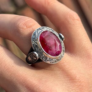 Peut inclure: Bague en argent ornée d'une pierre précieuse rouge foncé de forme ovale. La bague présente un motif gravé et ornemental autour de la pierre et sur le corps. Un bijou de caractère, idéal pour les occasions spéciales.