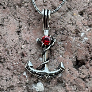 Peut inclure: Un collier avec un pendentif en forme d'ancre en argent avec une pierre précieuse rouge. L'ancre est ornée d'une corde et d'une croix.