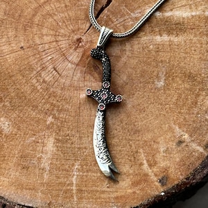 Może przedstawiać: Srebrny naszyjnik z zawieszką w kształcie miecza. Miecz jest ozdobiony czarnymi i czerwonymi kamieniami. Zawieszka jest grawerowana arabską kaligrafią.