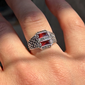 Puede incluir: Un anillo plateado con un diseño rectangular con tres piedras rojas y pequeños detalles redondos plateados. El anillo tiene un diseño de nudo celta en los laterales.
