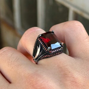 May include: A silver ring with a large, rectangular, deep red gemstone. The ring has a detailed design with smaller red stones around the main stone and decorative patterns on the band. The ring is shown on a finger.