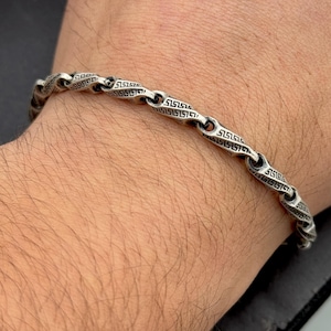 Könnte beinhalten: Ein silbernes Armband mit einem Kettenglieddesign, das ein griechisches Schlüsselmuster aufweist.
