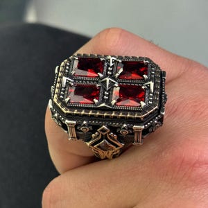 Könnte beinhalten: Silberfarbener Ring mit rechteckiger Fassung, besetzt mit vier quadratischen, tiefroten Edelsteinen. Der Ring hat aufwendige Details mit goldenen Akzenten und ein dekoratives Design am Band.