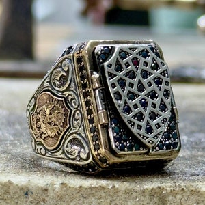 Puede incluir: Un anillo de plata con un diseño detallado que presenta un escudo con piedras preciosas negras y una cresta dorada. El anillo tiene una forma cuadrada y está engastado con piedras preciosas negras alrededor de los bordes.