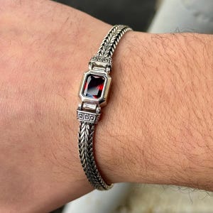 May include: Silver bracelet with a woven chain design. The bracelet features a rectangular, deep red gemstone set in a silver bezel. The bracelet is worn on a wrist.