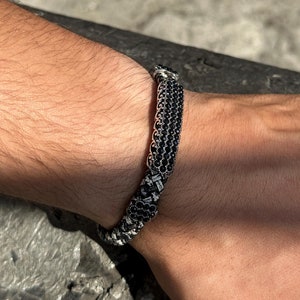 May include: A black and white braided bracelet with a silver clasp. The bracelet is made of small beads and is worn on a person's wrist.