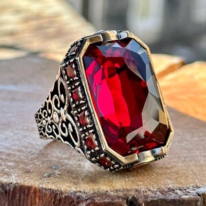 May include: A silver ring with an ornate design features a large, faceted, red gemstone.