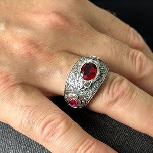 Peut inclure: Une bague en argent avec un motif orn&eacute; et une grosse pierre pr&eacute;cieuse rouge. La bague est port&eacute;e sur un doigt.
