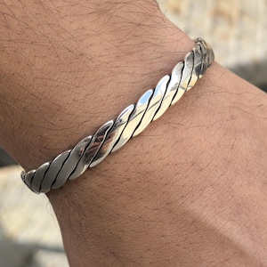Puede incluir: Un brazalete de plata con un diseño trenzado. El brazalete está hecho de metal y tiene un acabado pulido.