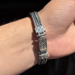 Könnte beinhalten: Ein silbernes Armband mit einem detaillierten, verzierten Verschluss. Das Armband besteht aus einem geflochtenen Kettenmuster.