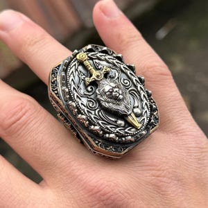 以下が含まれることがあります： ライオンの頭と剣を特徴とする詳細なデザインのシルバーリング。リングは、テクスチャ加工された縁のある正方形のベゼルにセットされています。