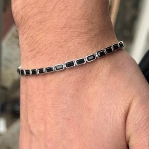 May include: A silver bracelet with rectangular black stones. The bracelet is worn on a wrist, showcasing the jewelry's design and color contrast. The bracelet is a stylish accessory.