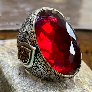 Può includere: Un anello d'argento con un designo ornato e una grande pietra rossa sfaccettata.