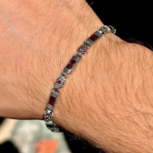 Può includere: Un bracciale in argento con pietre preziose rosse rettangolari e dettagli ornati. Il bracciale è indossato su un polso, mettendo in risalto il suo design. Le pietre sono di un rosso intenso, in contrasto con l'argento. Lunghezza bracciale: circa 19 cm.