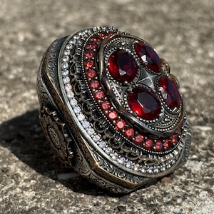 May include: An ornate silver ring with a large, oval, red gemstone at the center, surrounded by smaller red and clear stones. The ring has intricate detailing and a vintage aesthetic. The ring is set against a textured, gray surface.