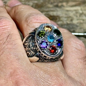 Può includere: Un anello d'argento con una faccia ovale incastonata con sette pietre preziose colorate. Le pietre sono disposte in un motivo circolare, con colori tra cui rosso, blu, verde e giallo. L'anello ha un design ornato e inciso sulla fascia.