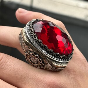 May include: A silver ring featuring a large, red, faceted gemstone set in an intricate, ornate silver setting.