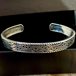 May include: Silver-toned cuff bracelet with an intricate, repeating wave and swirl pattern. The open-ended bracelet is displayed inside a black box, showcasing the detailed design and metallic finish. A classic piece of jewelry.