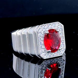 Op de afbeelding: Een zilveren ring met een grote, ovaal geslepen, helderrode edelsteen in het midden, omringd door een halo van kleinere, heldere stenen. De ring heeft een rechthoekige kop en een gepolijste band.