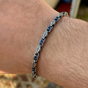 May include: Silver bracelet with ornate detailing and small, round blue gemstones. The bracelet is worn on a wrist, showcasing its design and craftsmanship. The links have an intricate pattern, creating a classic and elegant look.