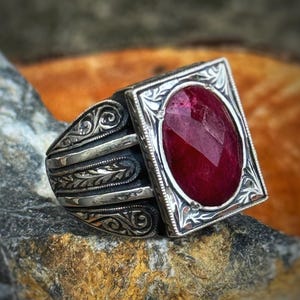 May include: A silver ring with an ornate design and a large, faceted, oval-shaped ruby gemstone. The ring's setting is square with decorative edges. The ring is displayed on a textured, gray stone surface.