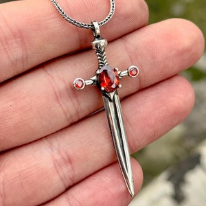 Könnte beinhalten: Ein silberner Schwertanhänger mit einem roten Edelstein in der Mitte und drei kleineren roten Edelsteinen an den Seiten. Der Anhänger ist an einer silbernen Kette.