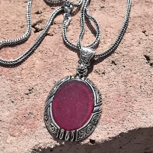 May include: A silver necklace with an oval pendant. The pendant has a deep red, oval-shaped stone set in an ornate, filigree-style frame. The chain is a textured, snake-style design.