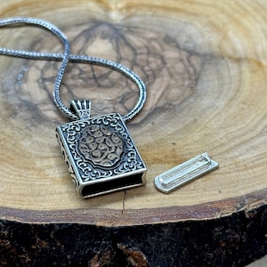 May include: A silver necklace with a detailed, ornate pendant shaped like a book. The pendant is open, revealing a smaller silver piece inside. The necklace is on a wooden surface.