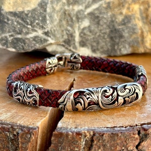 Pode incluir: Um bracelete de couro vermelho com detalhes decorativos prateados. O bracelete apresenta uma banda de couro trançada e um fecho prateado.