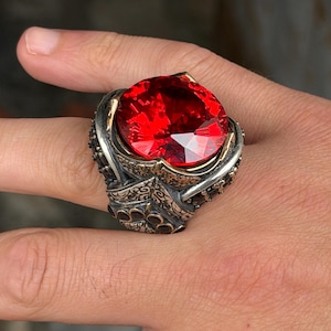 May include: A large silver ring featuring a prominent, round, faceted red gemstone set in a detailed, oxidised metal band with intricate, embossed patterns and dark recessed areas surrounding the central stone, worn on a finger.