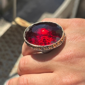 May include: A silver ring with an ornate design and a large, oval-shaped red gemstone.