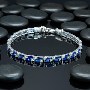 May include: A silver bracelet with alternating blue and clear rectangular stones. The bracelet is set against a black reflective surface with black stones in the background. The bracelet is a piece of jewelry.