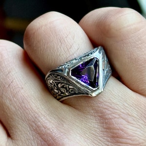 May include: A silver ring featuring an elaborate floral design and a large, triangular, purple gemstone.