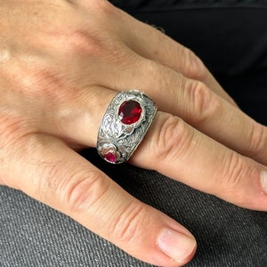 Peut inclure: Une bague en argent avec un design complexe et trois pierres pr&eacute;cieuses rouges. La bague est port&eacute;e au doigt d'une personne.