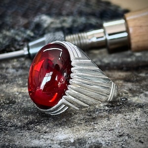 May include: A silver ring featuring a large, oval, deep red gemstone. The ring has a detailed, textured design on the sides. The gemstone reflects light, creating a vibrant appearance. The ring is set against a textured, grey surface.