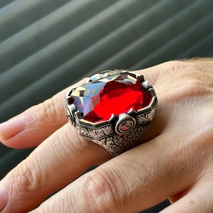 Peut inclure: Une bague en argent avec un motif orné mettant en vedette une grande pierre précieuse rouge. La bague présente un motif floral détaillé et une plus petite pierre précieuse claire.