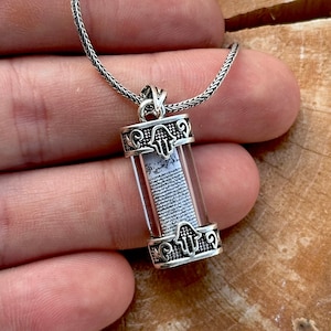 May include: Silver necklace featuring a clear glass pendant enclosing a miniature scroll. The pendant is adorned with a Hamsa hand charm.