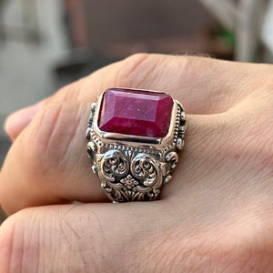 May include: A silver ring featuring a large, rectangular, deep red gemstone. The ring has an ornate, detailed design with swirling patterns and floral accents around the gemstone setting. The ring is on a finger.