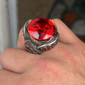 May include: A silver ring featuring a large, round, faceted red gemstone set in a decorative, oxidised metal band. The band displays intricate, textured patterns and is accented with smaller, embedded red stones along the sides of the setting.