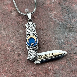 Könnte beinhalten: Silberne Anhänger Halskette mit einem messerförmigen Anhänger. Der Anhänger hat ein blaues und goldenes Halbmond- und Stern-Design. Die Messerklinge hat goldene Filigranarbeiten. Die Halskette ist aus Silber.