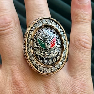 Könnte beinhalten: Ein verzierter goldfarbener Ring mit ovaler Fassung. Die Fassung zeigt ein zentrales Emblem mit grünen, roten und weißen Akzenten, umgeben von kleinen, klaren Steinen. Der Ring hat ein detailliertes, gemustertes Band.