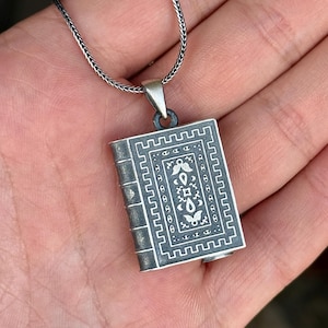 May include: A silver pendant in the shape of a book, with intricate engraved details. The pendant hangs from a silver chain. The book design features a detailed border and decorative elements. The pendant is held in a person's hand.