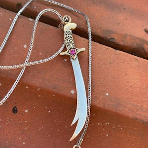 Könnte beinhalten: Eine silberne Kettenkette mit einem goldenen und silbernen Anhänger in Form eines gekrümmten Schwertes mit einem rosafarbenen Edelstein am Griff.