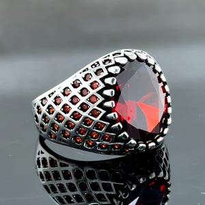 Peut inclure: Bague argentée ornée d'une grosse pierre rouge foncé facettée. Le corps de la bague présente un motif en treillis, avec de petites pierres rouges incrustées. La pierre rouge est le point central.